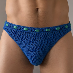 May include: A blue crochet brief featuring a textured, open-knit pattern throughout the main body. The waistband is accented with a woven, vibrant green ribbon threaded through the crochet stitches, creating a repeating decorative detail.