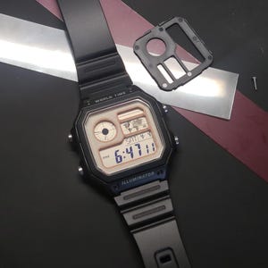 May include: Black digital watch with a rectangular face displaying the time 6:47:11 PM, Saturday, and a world map icon. The watch has a black strap and the text "WORLD TIME" and "ILLUMINATOR" are visible. A black watch component and a screw are also present.