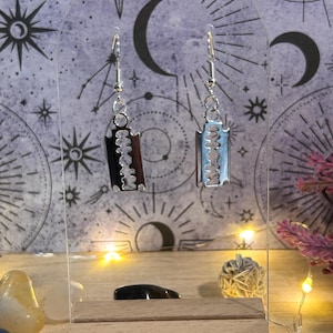 May include: A pair of silver-toned dangle earrings. Each earring features a razor blade design, one in black and brown, the other in light blue and silver. The earrings are displayed on a clear stand against a celestial-themed backdrop.