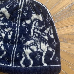 May include: A navy blue knit beanie with a white geometric pattern. The hat features a repeating design of figures and star-like shapes. The brim and top of the hat are solid navy blue.