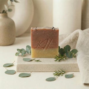May include: A rectangular bar of handmade soap with a two-tone color scheme: a light yellow base and a reddish-brown top. The soap is decorated with an engraved design and surrounded by green eucalyptus leaves and sprigs.