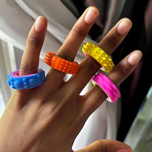 May include: A hand displays five colourful rings. The rings are blue, orange, yellow, and pink. Each ring has a unique textured design, including dots and spiky patterns. The rings are made of a flexible material.