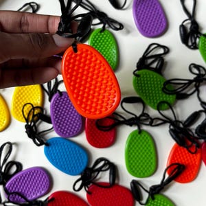Peut inclure: Une collection de pendentifs sensoriels de forme ovale présentant une surface texturée et bosselée sur une face. Chaque pendentif est attaché à un cordon noir avec un fermoir réglable. Les pendentifs sont disponibles en plusieurs couleurs vives, notamment orange, vert, bleu, violet, rouge et jaune.