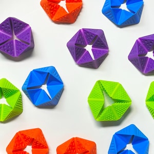 Könnte beinhalten: Eine Sammlung von mehrfarbigen, geometrischen, sechseckigen sensorischen Fidget-Spielzeugen aus Silikon. Jedes Teil verfügt über eine zentrale Öffnung und verschiedene strukturierte Oberflächen, darunter erhabene Punkte, diagonale Linien und kleine Dreiecke in Orange, Blau, Lila und Grün.