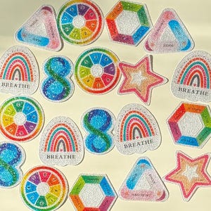 Op de afbeelding: Een verzameling kleurrijke, geborduurde patches in verschillende vormen: cirkels, regenbogen, sterren en zeshoeken. Sommige patches bevatten het woord "Breathe" of cijfers. Het kleurenpalet omvat regenboogtinten en pasteltinten.