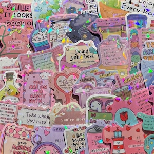 Peut inclure: Une collection d'autocollants holographiques présentant divers motifs aux tons pastel, incluant des citations motivantes, des illustrations de tasses à café, de fleurs, de cœurs et d'objets du quotidien, avec une finition scintillante.