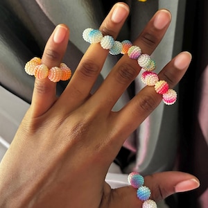 May include: Three rings made of small, textured, spherical beads in a gradient of rainbow colours. The beads transition from white to yellow, orange, pink, blue, and purple hues. Each ring features a flexible, beaded band design that encircles the finger.