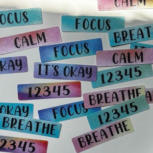 Op de afbeelding: Een verzameling rechthoekige glitterstickers in diverse kleurverlopen met zwarte tekst die FOCUS, CALM, IT'S OKAY, BREATHE en 12345 leest. De stickers zijn verspreid gerangschikt en tonen verschillende kleurencombinaties.