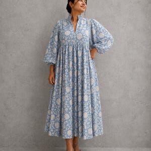 May include: A midi-length dress in a light blue and white floral print. The garment features a high collar with a V-neck opening, long voluminous puff sleeves with gathered cuffs, and a gathered waistline that flows into a full, pleated skirt.