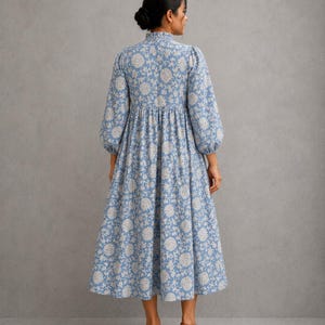 May include: A midi-length dress in a dusty blue hue featuring a cream-colored floral pattern. The garment has a high, ruffled collar, long sleeves with gathered cuffs, and a gathered waistline that flows into a full, pleated skirt silhouette.