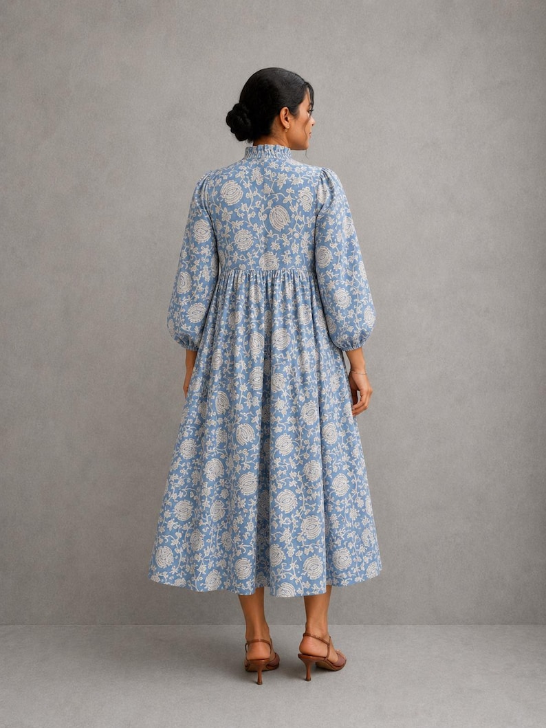 May include: A midi-length dress in a dusty blue hue featuring a cream-colored floral pattern. The garment has a high, ruffled collar, long sleeves with gathered cuffs, and a gathered waistline that flows into a full, pleated skirt silhouette.
