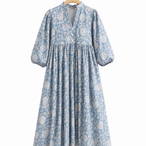 May include: A midi-length dress in light blue with a white floral pattern. It features a V-neckline with three white buttons, a gathered waistline, and three-quarter length puff sleeves with elasticated cuffs, hanging on a wooden hanger.