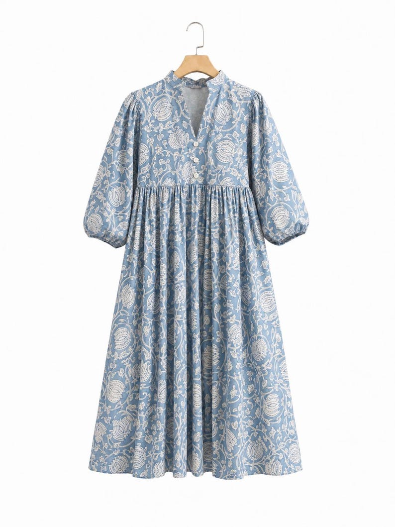 May include: A midi-length dress in light blue with a white floral pattern. It features a V-neckline with three white buttons, a gathered waistline, and three-quarter length puff sleeves with elasticated cuffs, hanging on a wooden hanger.