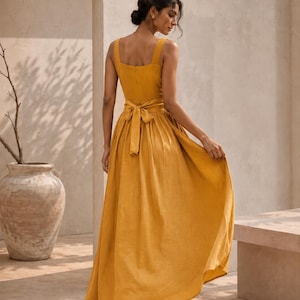 Può includere: Un abito lungo senza maniche color giallo senape con scollo quadrato, corpetto aderente e gonna ampia e fluida. L'abito include una cintura in tessuto abbinata in vita, annodata con un fiocco sul retro, e una chiusura con zip nascosta.