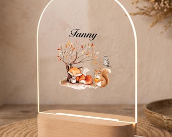 May include: An arched clear acrylic night light with a wooden base featuring a printed illustration of a fox, a squirrel, and an owl under a tree with the name Fanny written in black script above the animals and a small orange pumpkin.