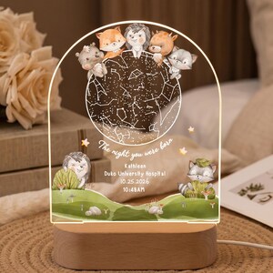 以下が含まれることがあります： 木製ベース付きのアーチ型アクリル製ナイトライト。星図と「The night you were born, Kathleen, Duko University Hospital, 10.25.2026, 10:48AM」というテキストが描かれています。デザインにはキツネ、ハリネズミ、シカ、アライグマのイラストが含まれています。