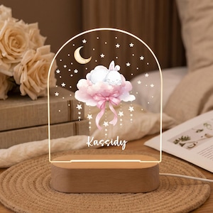 May include: An arched acrylic night light with a wooden base featuring a white bunny sleeping on a pink cloud, a yellow crescent moon, and white stars. The name Kassidy is printed in white script below the cloud, with dangling star accents.