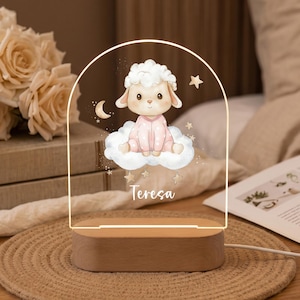 Puede incluir: Una luz nocturna de acrílico en forma de arco con base de madera que presenta una ilustración de un cordero en pijama rosa sentado sobre una nube, rodeado de estrellas y una luna creciente, con el nombre Teresa escrito en blanco abajo.