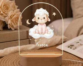 May include: An arched acrylic night light with a wooden base featuring a printed illustration of a lamb in pink pajamas sitting on a cloud, surrounded by stars and a crescent moon, with the name Teresa printed in white script at the bottom.