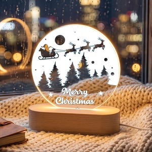 May include: A circular acrylic night light with a wooden base featuring a black silhouette of Santa Claus in a sleigh pulled by three reindeer flying over a forest of pine trees. The text Merry Christmas is printed in white in the center.