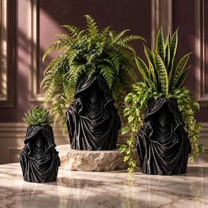 May include: Three black planters shaped like hooded figures in varying sizes. The smallest holds a succulent, the middle one contains a large fern, and the largest features a snake plant and trailing pothos vines, all arranged on a stone base.