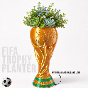 May include: A gold-colored trophy-shaped planter featuring a globe design at the top and a green base with the text FIFA WORLD CUP. The planter contains various green succulents and small blue and white flowers. Text reads FIFA TROPHY PLANTER.