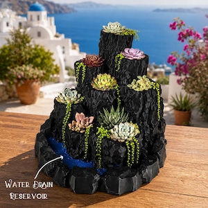 May include: A tiered black planter with a textured, rock-like surface featuring multiple succulent plants and trailing greenery. The structure includes a blue water drain reservoir at the base with white text pointing to it labeled Water Drain Reservoir.