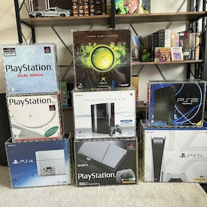 May include: A collection of video game consoles, including PlayStation, Xbox, and PS2, PS4, and PS5. The consoles are in their original boxes, stacked together. The boxes are various colors, with the PlayStation boxes featuring the PlayStation logo.