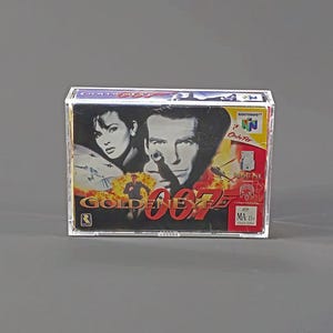 May include: A GoldenEye 007 Nintendo 64 video game cartridge in a clear plastic case. The game box features images of a man and woman, with the title in red and gold. The Nintendo 64 logo is visible.