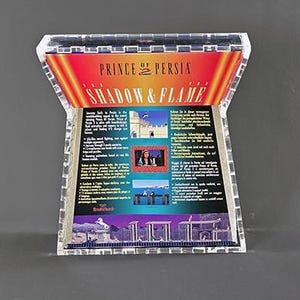 May include: A clear, acrylic display case featuring the game "Prince of Persia: The Shadow & Flame." The case has a colorful graphic with the game's title and imagery. The case is designed to showcase the game's packaging.