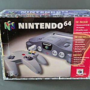 May include: A Nintendo 64 video game console box with a gray console and controller. The box features the Nintendo 64 logo and product details. The box is encased in a clear protective case.
