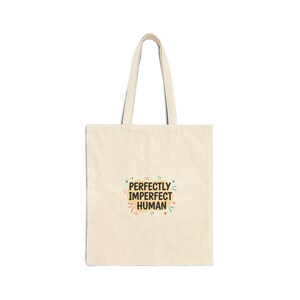 May include: A cream-colored tote bag with a pair of matching handles. The bag features the text "PERFECTLY IMPERFECT HUMAN" in a colorful, playful font. The design includes stars, dots, and a small leaf.