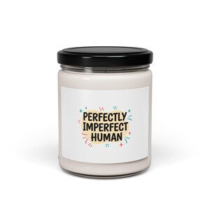 Puede incluir: Una vela blanca en un tarro de cristal con tapa negra. La etiqueta presenta el texto "PERFECTLY IMPERFECT HUMAN" en una fuente colorida y juguetona, con acentos de estrellas y remolinos. Es un artículo de decoración del hogar.