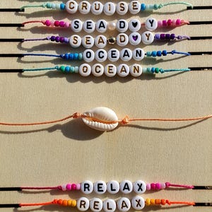 May include: A wooden display board with several beaded bracelets. The bracelets spell out words like "CRUISE", "SEA", "OCEAN", "RELAX", "SUN", and "SAIL". A seashell is also featured. The Duke's logo is at the top.