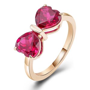 Op de afbeelding: Een goudkleurige ring met twee hartvormige, gefacetteerde, rode edelstenen. De edelstenen zijn naast elkaar geplaatst, verbonden door een kleine gouden staaf en gemonteerd op een gouden band. De ring is ontworpen om op een strik te lijken.