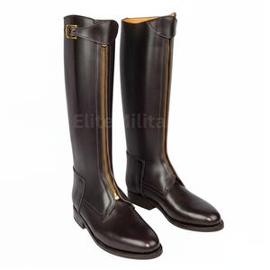 May include: A pair of dark brown leather riding boots with gold-coloured zips and buckles. The boots are tall, reaching the knee, and have a classic equestrian design. The text "Elite Milita" is visible on the boots.
