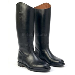 Puede incluir: Un par de botas altas de montar en cuero negro con acabado liso, que presentan una caña exterior alta, punta redondeada, tacón bajo y forro interior color canela. El texto EliteMilitaria es visible en el lateral de la caña de la bota.