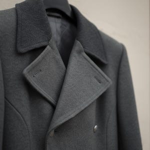 May include: A dark charcoal grey double-breasted coat featuring a wide notched lapel, a structured collar, and visible metallic buttons. The garment is crafted from a textured fabric and includes a smooth interior lining visible at the neckline.