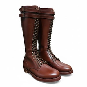 Puede incluir: Un par de botas altas de cuero marrón. Las botas presentan un diseño con cordones y múltiples ojales, además de dos correas con hebilla en la parte superior. Tienen una puntera redondeada y una suela resistente.