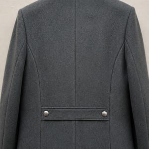 May include: A charcoal grey coat featuring a dark grey collar and a horizontal back strap detail. The strap is secured with two metallic silver buttons. The coat has a structured design with vertical seams running down the back panel.