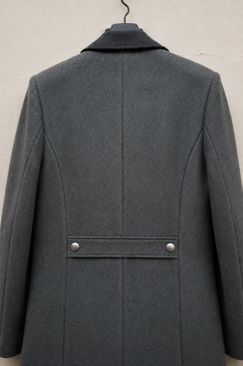 May include: A charcoal grey coat featuring a dark grey collar and a horizontal back strap detail. The strap is secured with two metallic silver buttons. The coat has a structured design with vertical seams running down the back panel.