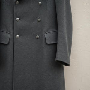 May include: A dark charcoal grey double-breasted coat featuring six round metallic buttons arranged in two vertical columns. The coat includes two horizontal flap pockets positioned at the waistline and long sleeves with clean, finished cuffs.