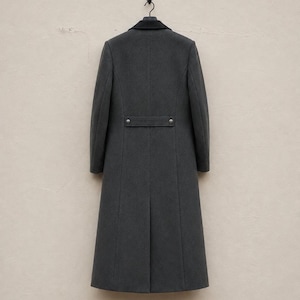 May include: A long, charcoal grey coat featuring a dark black collar and a horizontal back belt detail secured with two metallic buttons. The coat has a structured silhouette with vertical seams and a single vent at the bottom hem, hanging on a hanger.