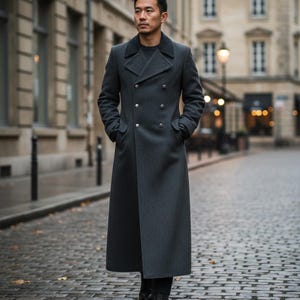 May include: A charcoal grey double-breasted long coat featuring a wide notched lapel, a dark collar, and two rows of silver-toned buttons. The coat extends to the lower calf and includes two side pockets, worn over a dark shirt and dark trousers.