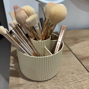 Puede incluir: Un soporte beige y acanalado para brochas de maquillaje lleno de varias brochas de maquillaje y una máscara de pestañas Lancôme. Las brochas tienen cerdas de color claro y mangos dorados. El soporte está sobre una superficie de madera.