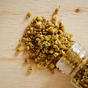 May include: Dried chamomile flowers spilling from a clear glass jar onto a light wooden surface. The flowers are yellow and brown, with small, round heads and delicate stems. The image is a top-down view.