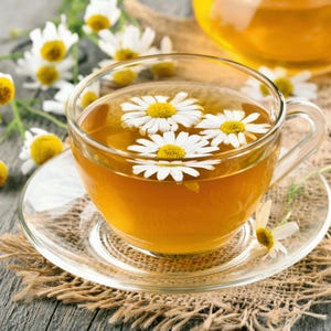 May include: A clear glass teacup and saucer filled with golden-colored tea and floating chamomile flowers. The cup sits on a burlap coaster, with more chamomile flowers and a teapot in the background. The scene evokes a sense of calm and wellness.