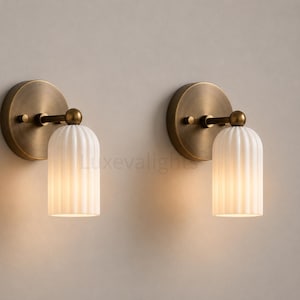 May include: A pair of wall sconces featuring circular antique brass backplates and short horizontal arms supporting cylindrical, vertically ribbed white glass shades that emit a soft, warm glow. The text Luxevalights is visible.