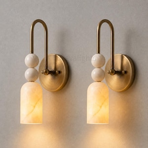 May include: Two wall sconces with a warm, inviting glow. Each sconce features a brass finish, a curved arm, and a cylindrical, off-white alabaster shade. The design includes three white marble spheres and a circular base.