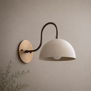 May include: A wall-mounted lamp featuring a circular brass backplate, a curved brass arm, and a matte off-white dome-shaped shade. The fixture includes a small light bulb visible inside the shade, with the text Luxvalights visible on the wall.